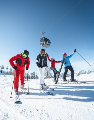Skiers rejoice and stand on the piste with a cable car hovering above them in the bright blue sky. | © Ski amadé