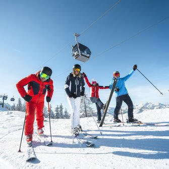 Skiers rejoice and stand on the piste with a cable car hovering above them in the bright blue sky. | © Ski amadé