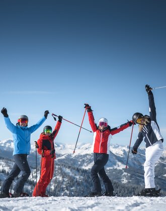 Four friends in ski equipment stand on the piste and stretch their arms joyfully in the air. | © Ski amadé