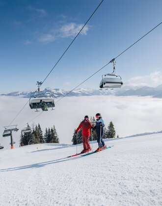 Zwei Skifahrer auf sonniger Piste unter Sessellift mit Blick auf Wolkenmeer und Berge in Ski amadé | © Ski amadé