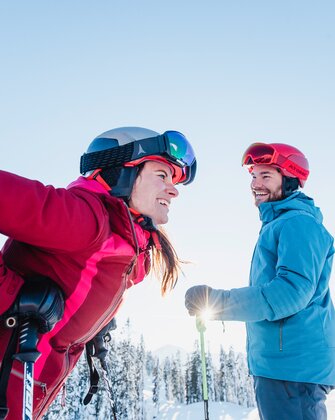 A female skier laughs and leans forward, while a male skier looks at her. | © Ski amadé