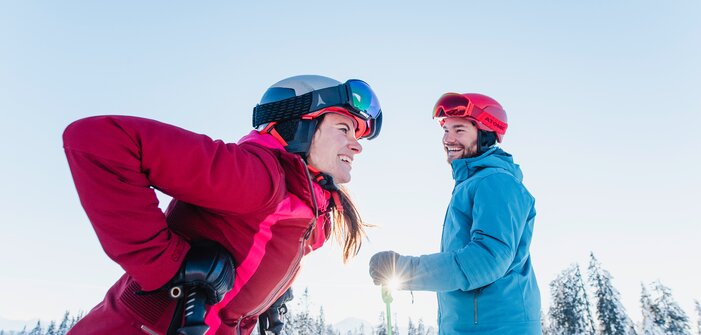 A female skier laughs and leans forward, while a male skier looks at her. | © Ski amadé