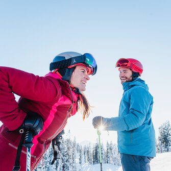 Eine Skifahrerin lacht und beugt sich vor, ein Skifahrer schaut sie an. | © Ski amadé