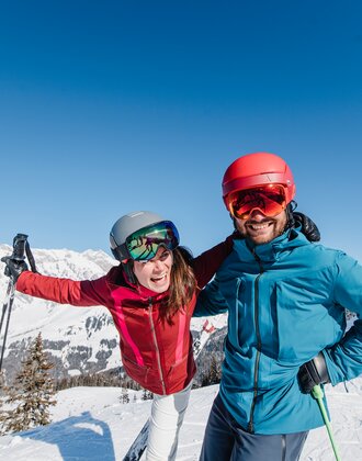 A smiling couple in ski gear poses in front of a snow-covered mountain landscape. | © Ski amadé