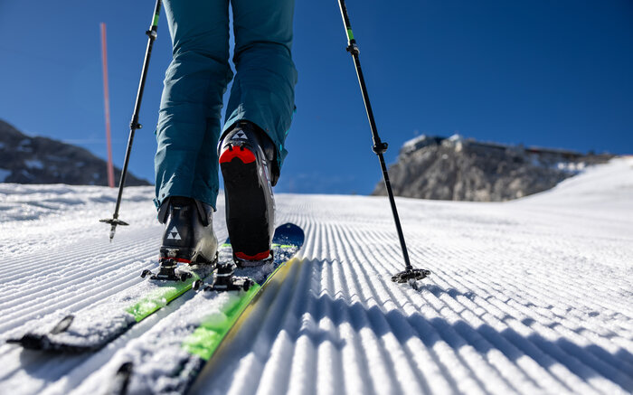 Close-up of a touring ski boot on groomed piste with poles and sunlit background | © Harald Steiner