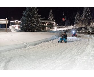 Familie rodelt bei Nacht auf beleuchteter Winterrodelbahn mit Stirnlampen in Gastein, im Hintergrund verschneite Hütte | © GTG_Marktl Photography