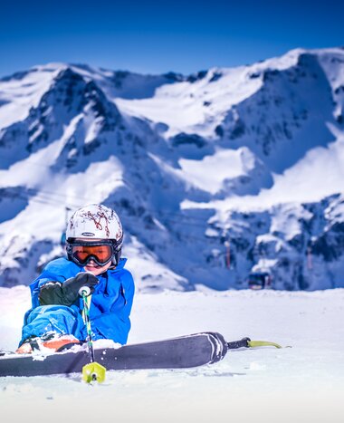 Kind in blauer Skibekleidung sitzt lachend im Schnee vor steilen Bergen in Gastein | © Max Steinbauer