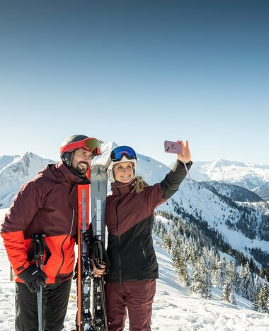 Couple in ski gear taking a selfie with smartphone and snowy mountain backdrop in Grossarltal | © TVB Großarltal - Lorenz Masser