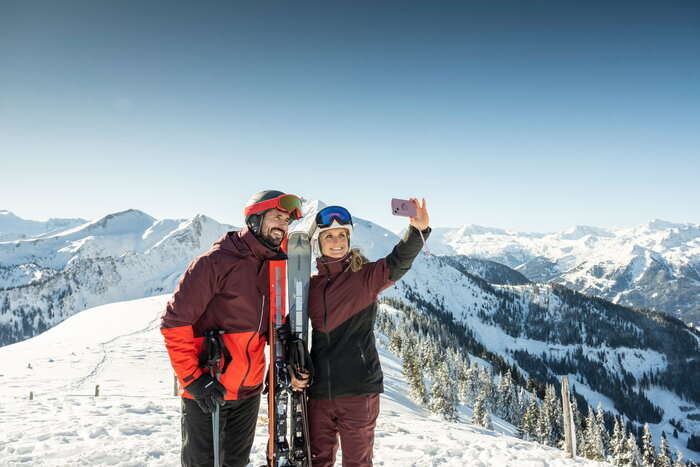 Couple in ski gear taking a selfie with smartphone and snowy mountain backdrop in Grossarltal | © TVB Großarltal - Lorenz Masser