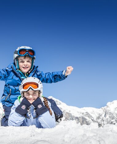 Zwei Kinder in Skikleidung spielen im Schnee vor verschneiten Alpenbergen | © Felsch Fotodesign