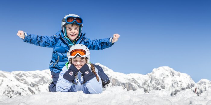 Zwei Kinder in Skikleidung spielen im Schnee vor verschneiten Alpenbergen | © Felsch Fotodesign