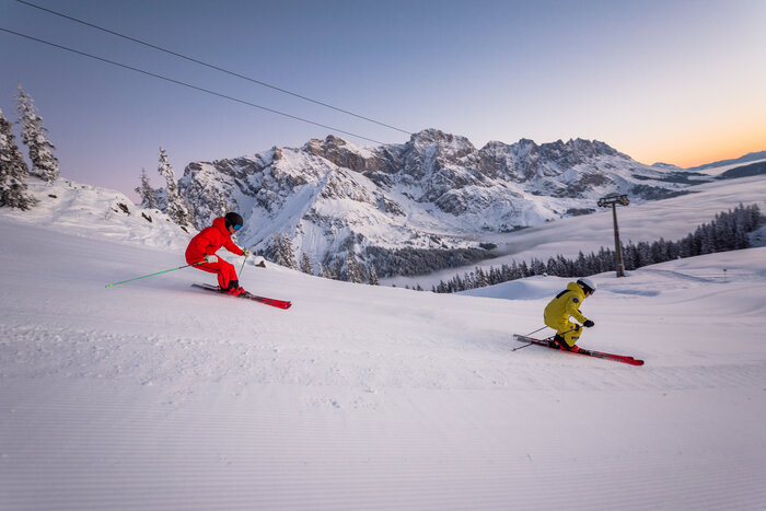 Ein Skifahrer in gelbem Skianzug und einer in rotem fahren die Piste hinunter und im Hintergrund geht die Sonne gerade auf | © Hochkönig Tourismus