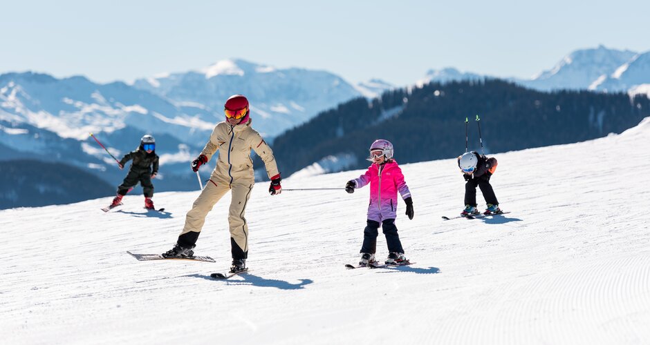 Erwachsene fährt mit drei Kindern auf breiter Piste bei Sonnenschein in den Alpen | © Hochkönig Tourismus GmbH