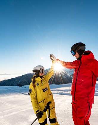 Two skiers in yellow and red gear give each other a high five on ski slope | © Hochkönig Tourismus GmbH