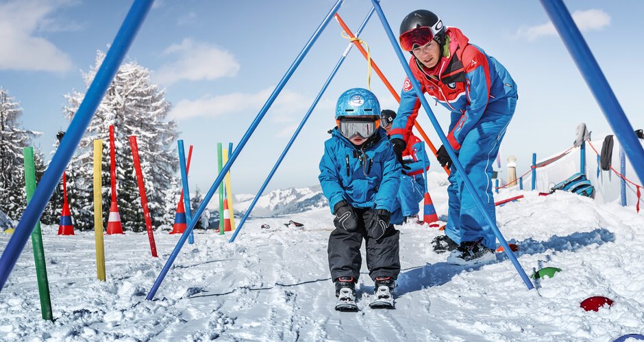 Slalom poles are stuck in the snow and tied together at the top and a child skis underneath while the ski instructor supervises everything