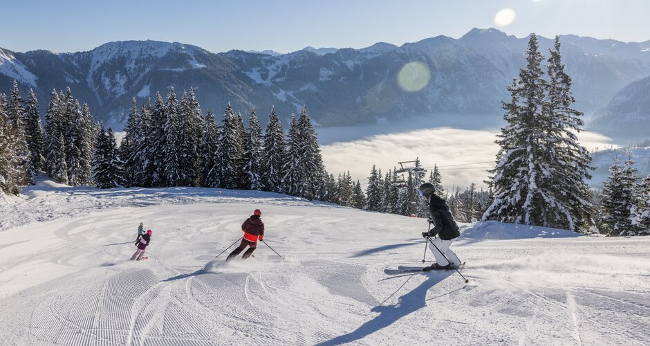 A family is skiing down the piste and the valley is covered in fog, while a bright blue sky can be seen above | © Michael Grössinger