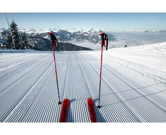 Two ski poles and red skis on freshly groomed ski slope with view of snow-covered alpine landscape | © Snow Space Salzburg Christian Schartner