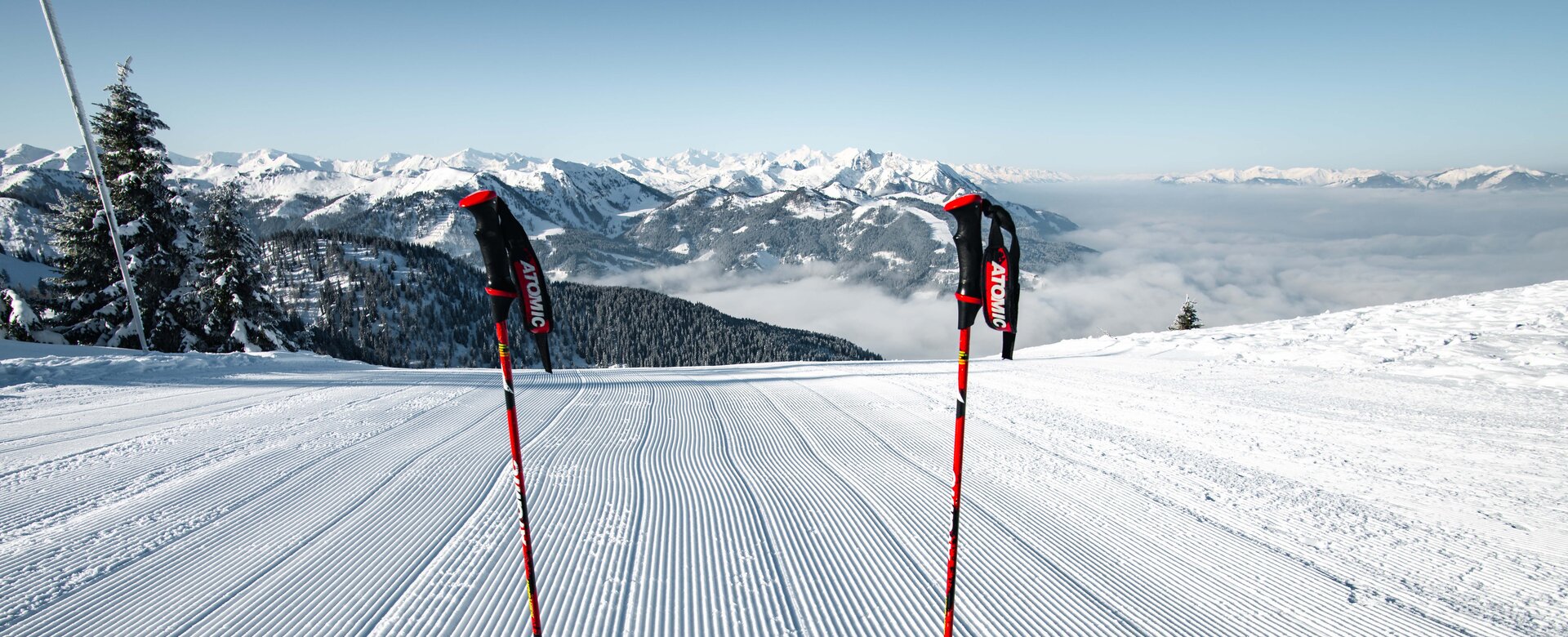 Two ski poles and red skis on freshly groomed ski slope with view of snow-covered alpine landscape | © Snow Space Salzburg Christian Schartner
