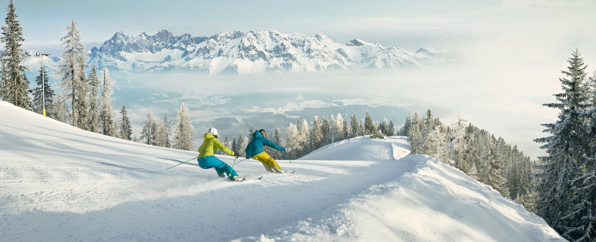 Zwei Skifahrer in bunter Kleidung fahren auf breiter Piste mit Blick auf verschneiten Dachstein | © Schladming-Dachstein - Peter Burgstaller