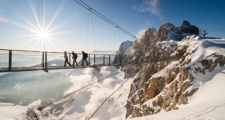 Vier Personen überqueren bei Sonne eine Hängebrücke am verschneiten Dachstein mit Blick ins Tal | © David McConaghy