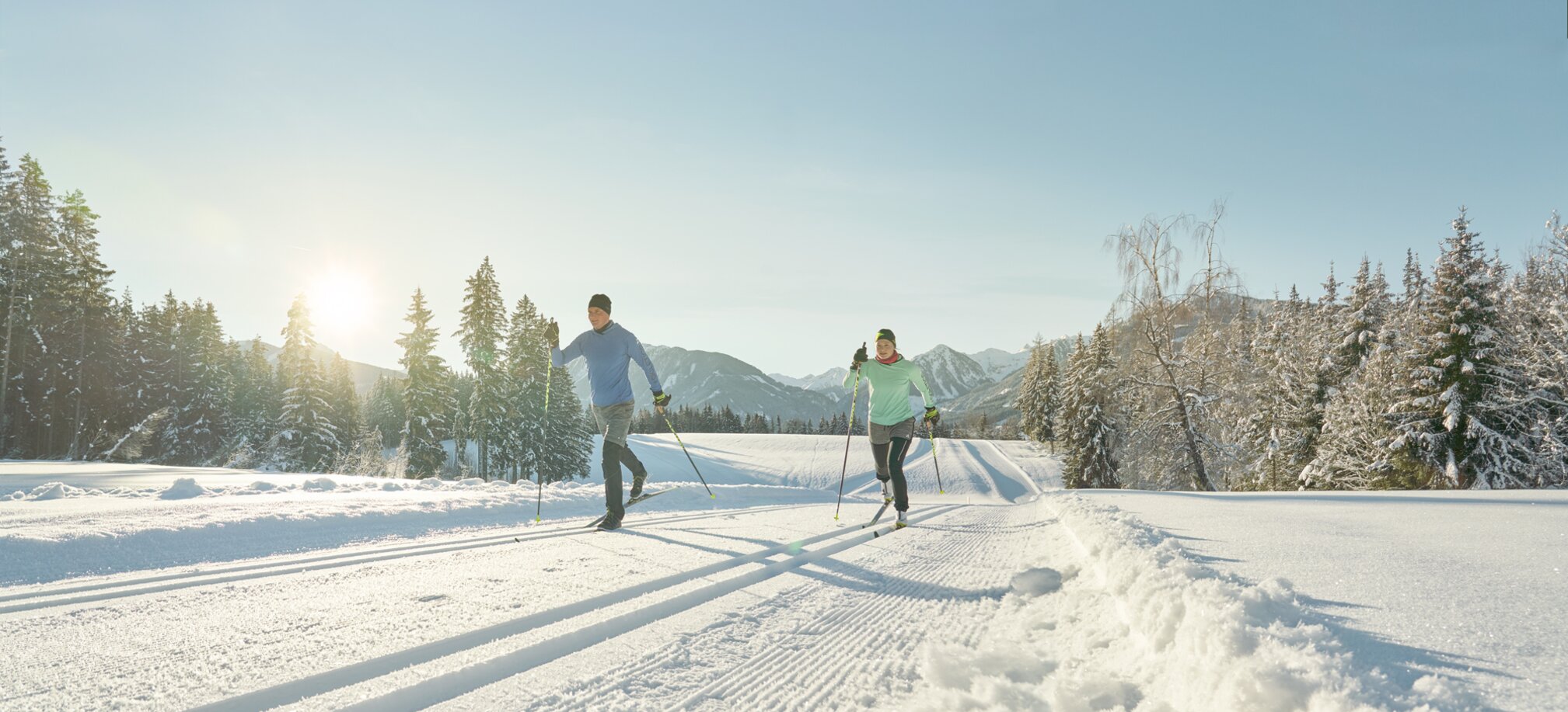 Zwei Langläufer in Sportkleidung gleiten bei Sonne auf gespurter Loipe durch verschneite Winterlandschaft | © Peter Burgstaller
