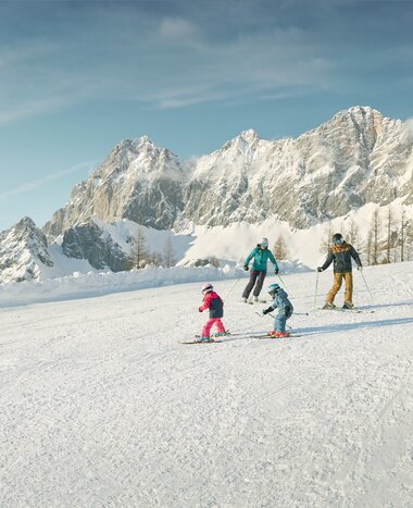 Zwei Erwachsene und zwei Kinder fahren Ski vor verschneitem Dachsteinmassiv in den Alpen | © Peter Burgstaller
