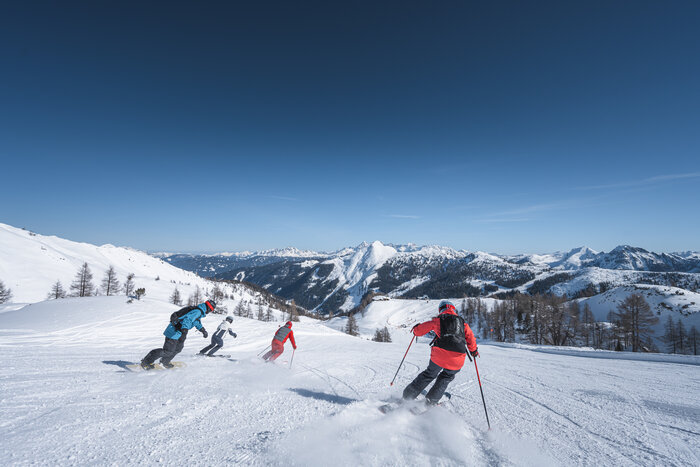 Drei Skifahrer und ein Snowboarder fahren gemeinsam talwärts auf Piste mit verschneiter Bergkulisse im Hintergrund.