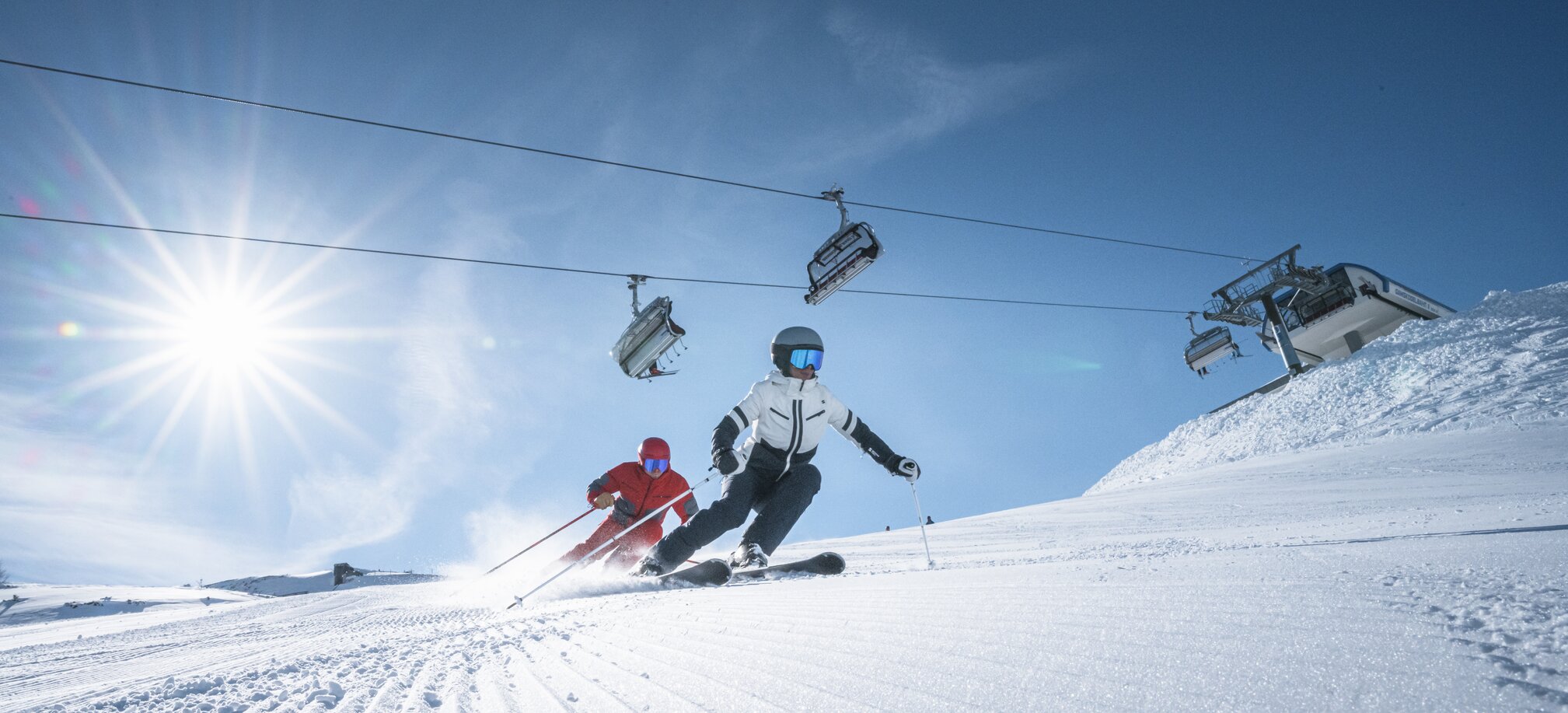 Two skiers ride side by side on groomed slope, above them a chairlift and bright sun shining through a clear sky.