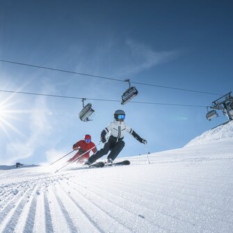Two skiers ride side by side on groomed slope, above them a chairlift and bright sun shining through a clear sky.