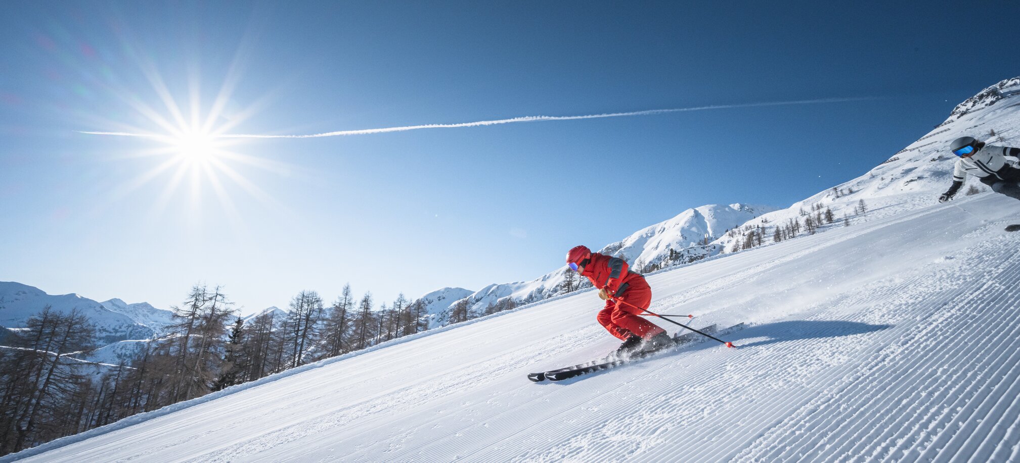Zwei Skifahrer carven bei strahlendem Sonnenschein über frisch präparierte Piste vor alpiner Winterkulisse.