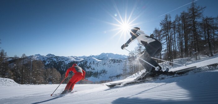 Two skiers carve down a freshly groomed slope, sunbeams shining through trees and above the mountain panorama.