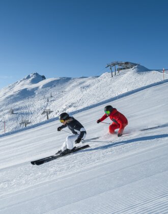 Skifahrer auf breiter Piste bei Sonne im Skigebiet der Alpen | © Two skiers skiing side by side on sunny groomed slope with view of snowy mountains and chairlift