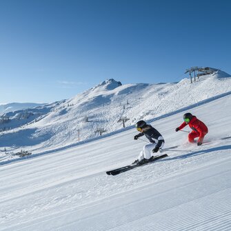 Two skiers carving gracefully on freshly groomed slopes with a stunning mountain backdrop in Ski amadé. Ideal conditions for unforgettable ski days.
