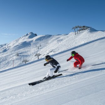Zwei Skifahrer fahren parallel auf sonniger, gewalzter Piste mit Blick auf verschneite Berge und Sessellift