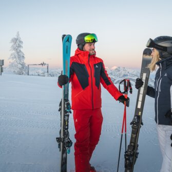 A man and a woman in ski gear stand with skis on a groomed slope in front of mountain scenery