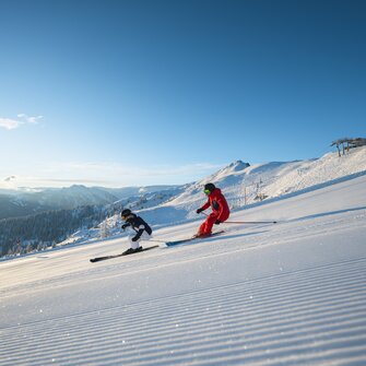 Zwei Skifahrer fahren parallel auf frisch präparierter Piste bei Sonne und Alpenpanorama im Hintergrund