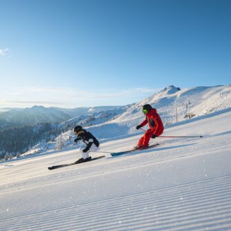 Two skiers skiing in parallel on a freshly groomed slope with sunshine and Alpine panorama in the background