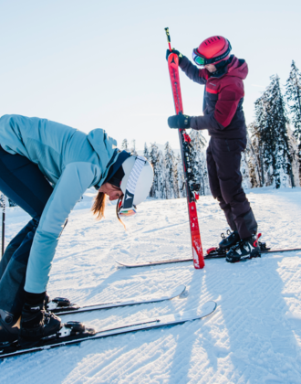 A skier bends down to her speedy skis by her ski boots and the skier is just looking at the unbuckled ski while he is still standing on the piste with the other one