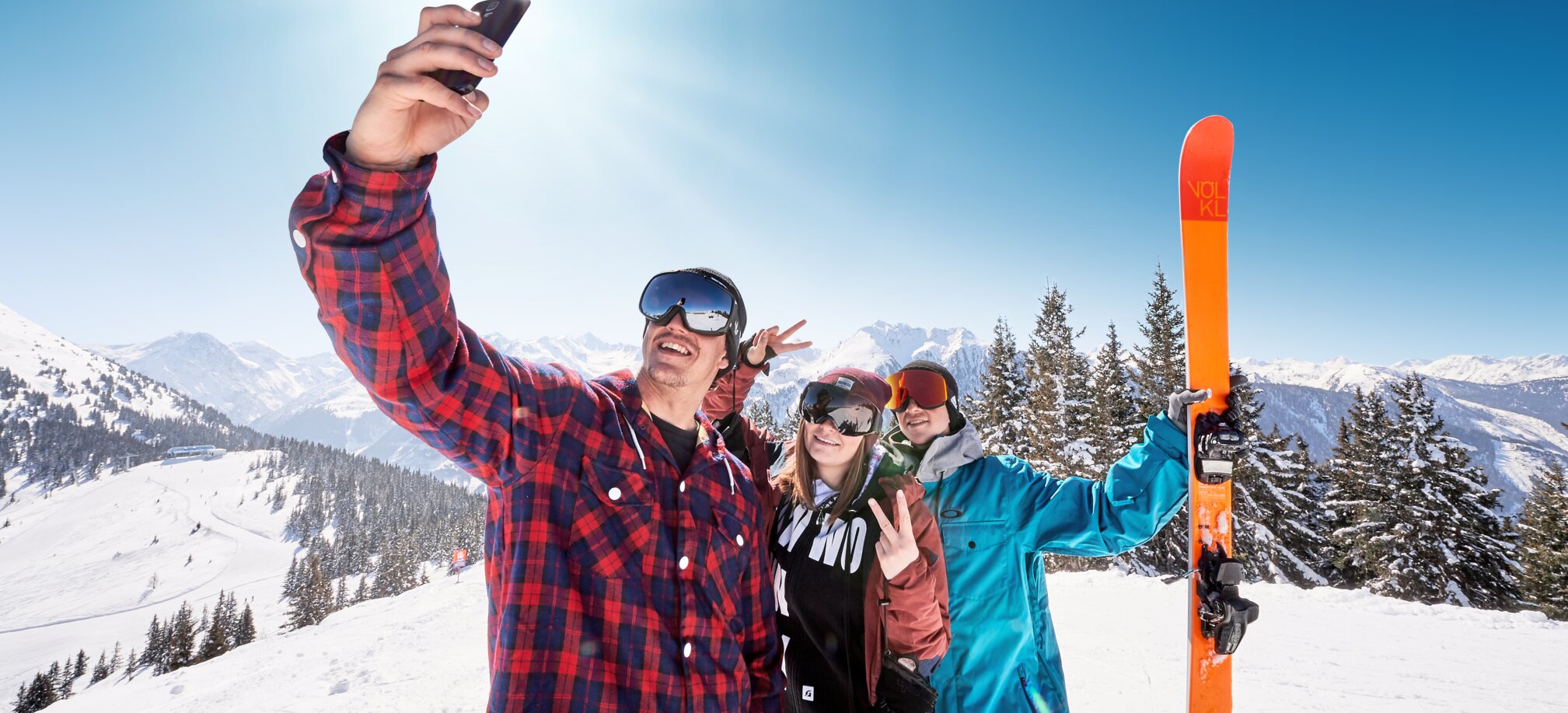 Three teenagers in ski goggles take a selfie on the snow-covered mountain and the one in the back holds an orange ski
