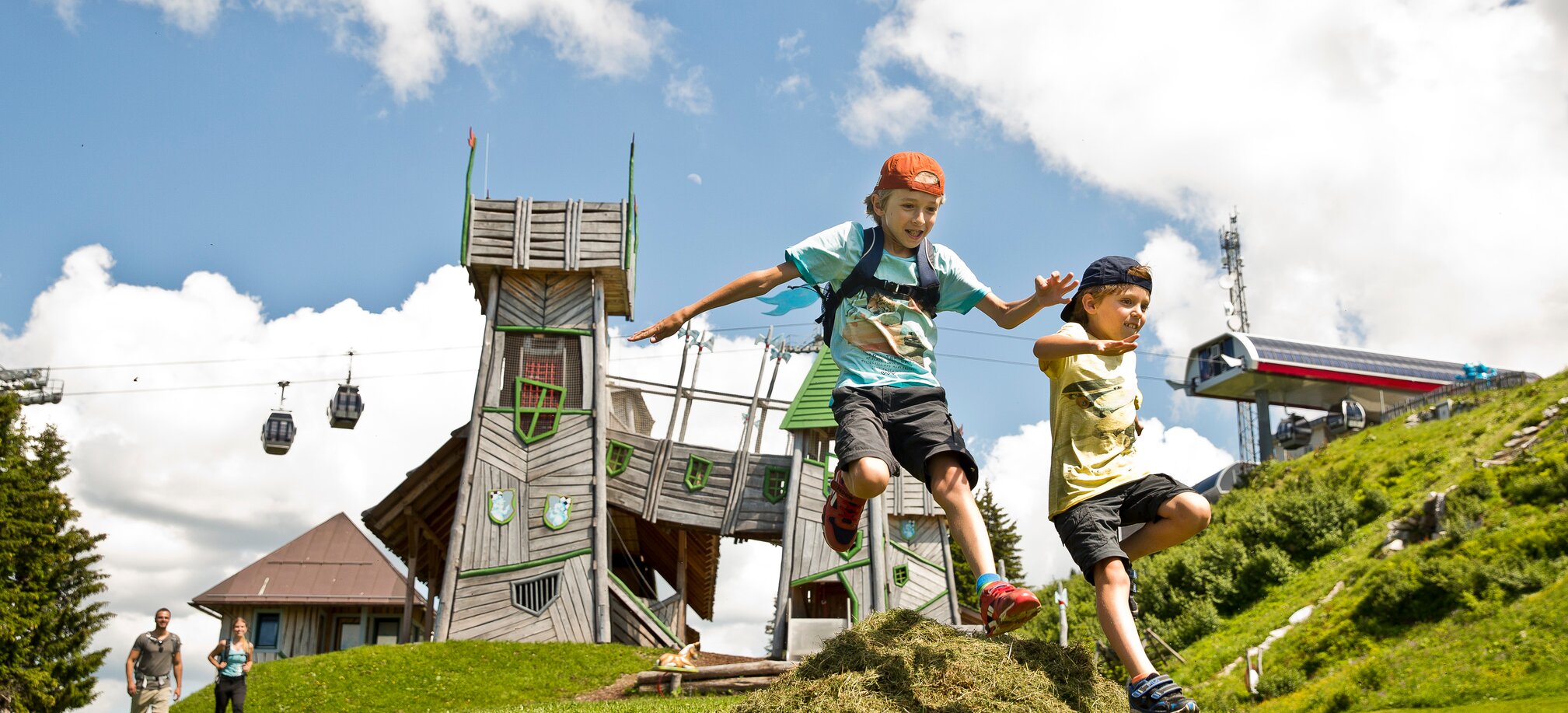 Two children are jumping over a small grassy hill and in the background you can see a playground that looks like a castle | © sanktjohann.com, Mirja Geh