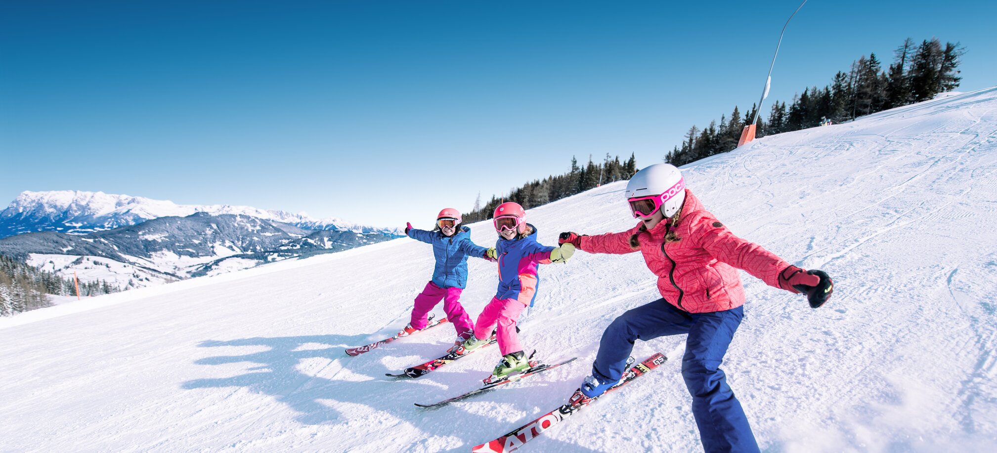 Drei Kinder halten sich an den Händen und fahren nebeneinander die Piste hinunter wobei sich die Skispitzen der einzelnen Kinder berühren und sie lachen dabei