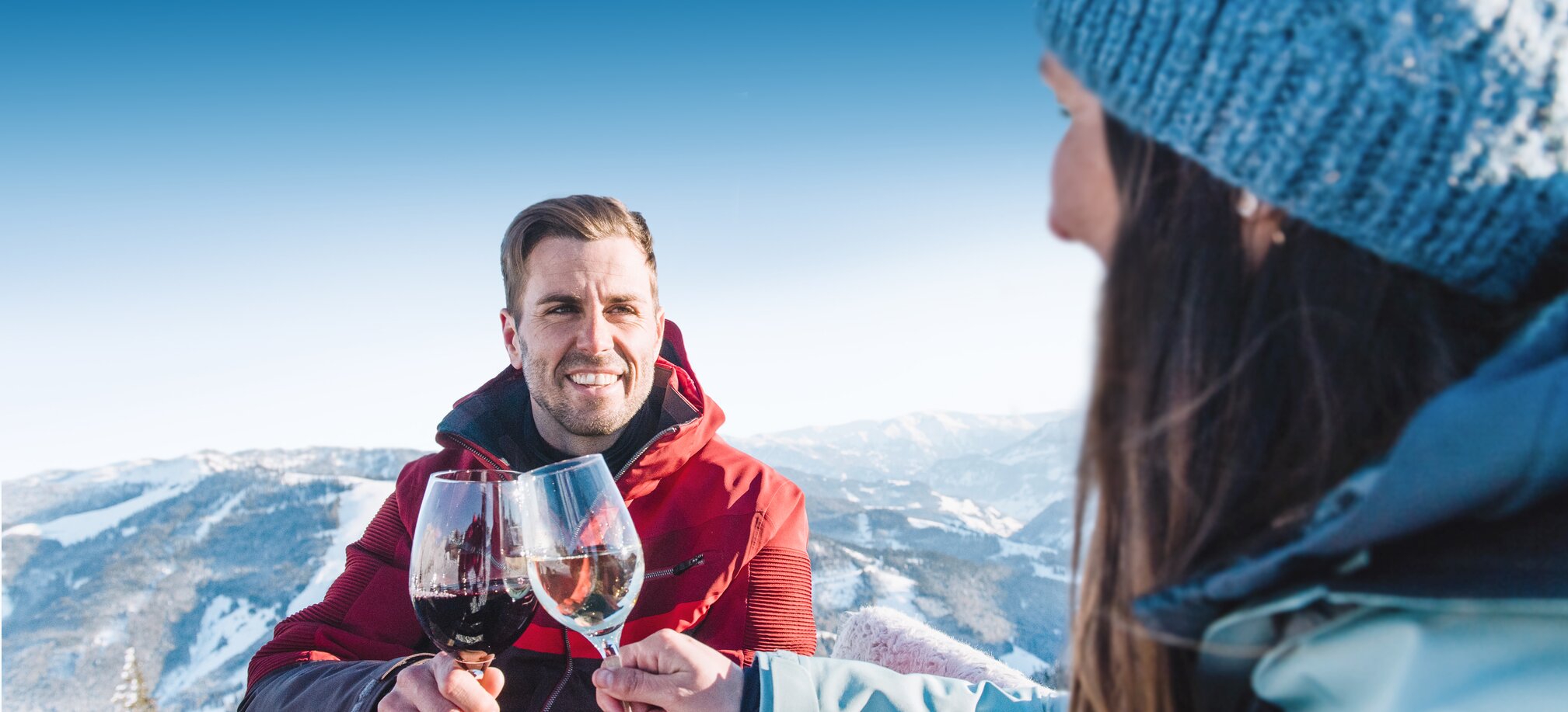 Ein Mann mit roter Skijacke lächelt und stoßt mit einem Weinglas mit einer Frau mit blauer Haube und blauer Skijacke an, während sie an einem Holztisch sitzen