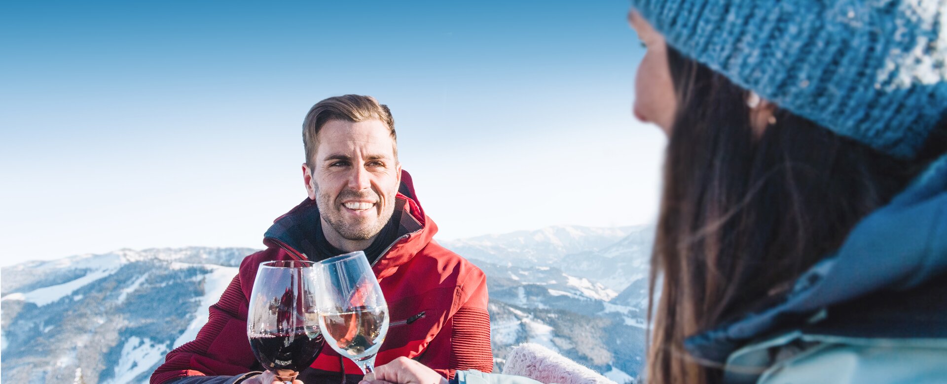 Ein Mann mit roter Skijacke lächelt und stoßt mit einem Weinglas mit einer Frau mit blauer Haube und blauer Skijacke an, während sie an einem Holztisch sitzen