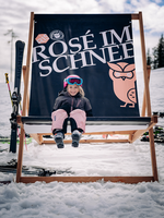 Lächelndes Kind mit Skibekleidung sitzt im Schnee auf großem Liegestuhl mit Banner Rose im Schnee