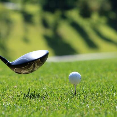 Ein Golfball steht auf einem Tee, kurz vor dem Abschlag mit einem Driver, im Hintergrund ein sonniges Green.
