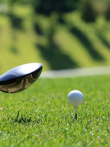A golf ball on a tee about to be hit with a driver club, surrounded by bright green grass on a sunny fairway.