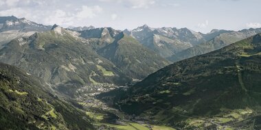 Wide view over Gastein Valley with a town below, green slopes and a long mountain range under a bright sky. | © Gasteinertal Tourismus, Marktl Photography