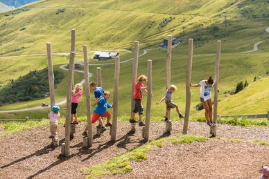 Mehrere Kinder balancieren auf Holzstämmen im Kinderareal der Schlossalm vor alpiner Sommerkulisse mit grünen Hängen | © Gasteiner Bergbahnen AG