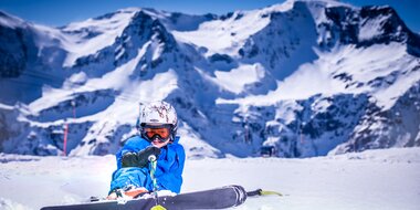 Child in blue ski gear sitting and laughing in snow with steep peaks in Gastein | © Max Steinbauer