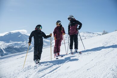Three people in ski gear are standing on a slope talking with smiles | © Gasteinertal Tourismus GmbH, Christoph Oberschneider